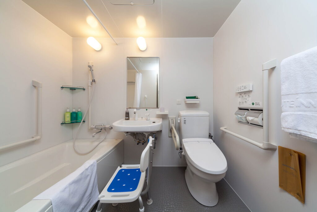 ルートインホテルズ、バリアフリールームのバス・トイレ設備の室内写真。浴槽や便器の周りには手すりがある。シャワーチェアも準備済
