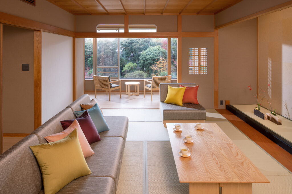 界 伊東のご当地部屋「伊豆花暦の間」の写真。畳の間の上にソファーとクッション、ローテーブルがある。奥には大きな窓があり外の景色を眺めることができる。